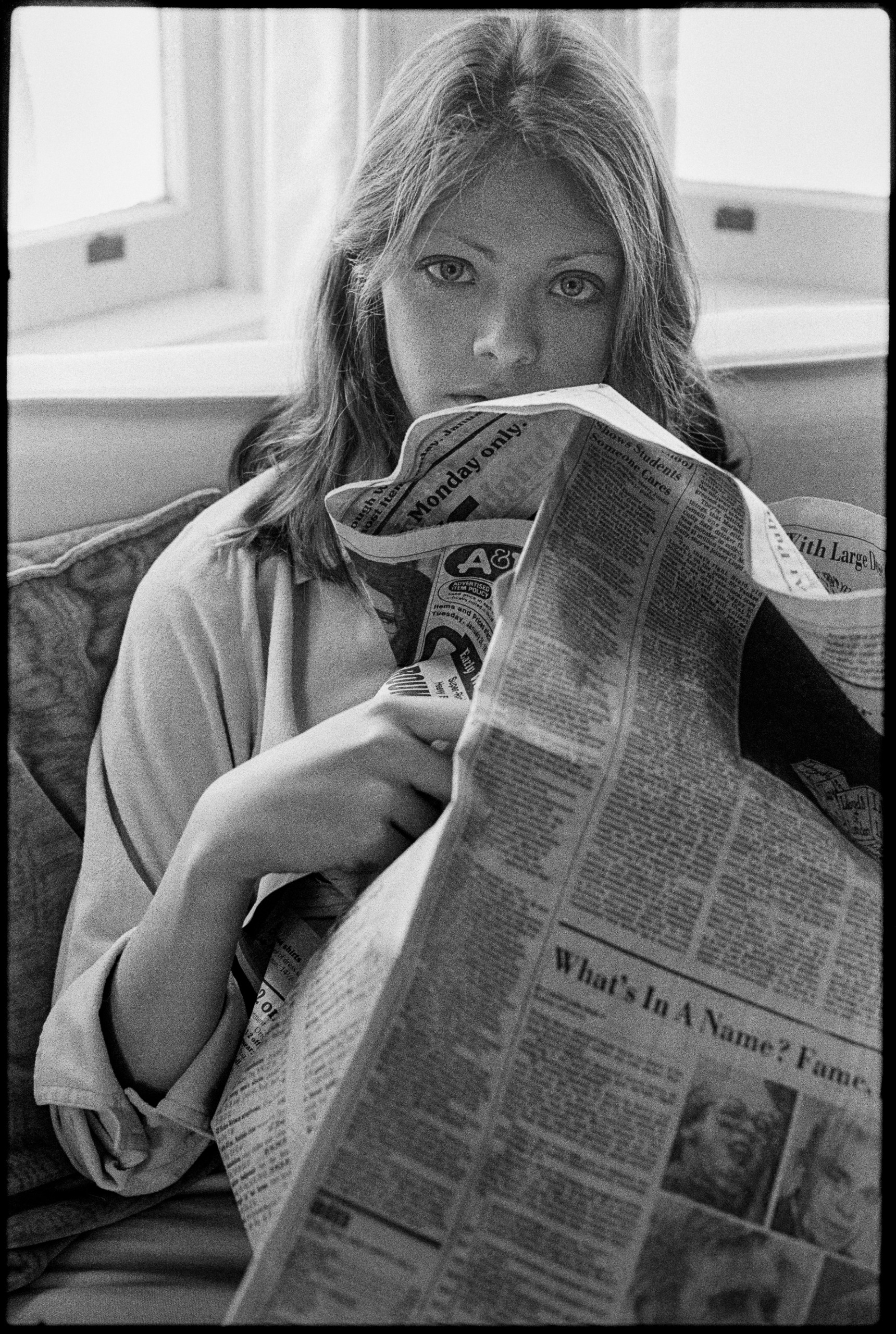 A black-and-white photo of a young woman looking over the top of a folded newspaper she is holding, with a window behind her.