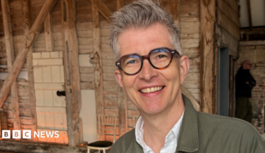 Gareth Malone smiles for the camera. He has short grey hair spiked up at the front, thick-rimmed glasses and an open-collar white shirt and khaki jean jacket.