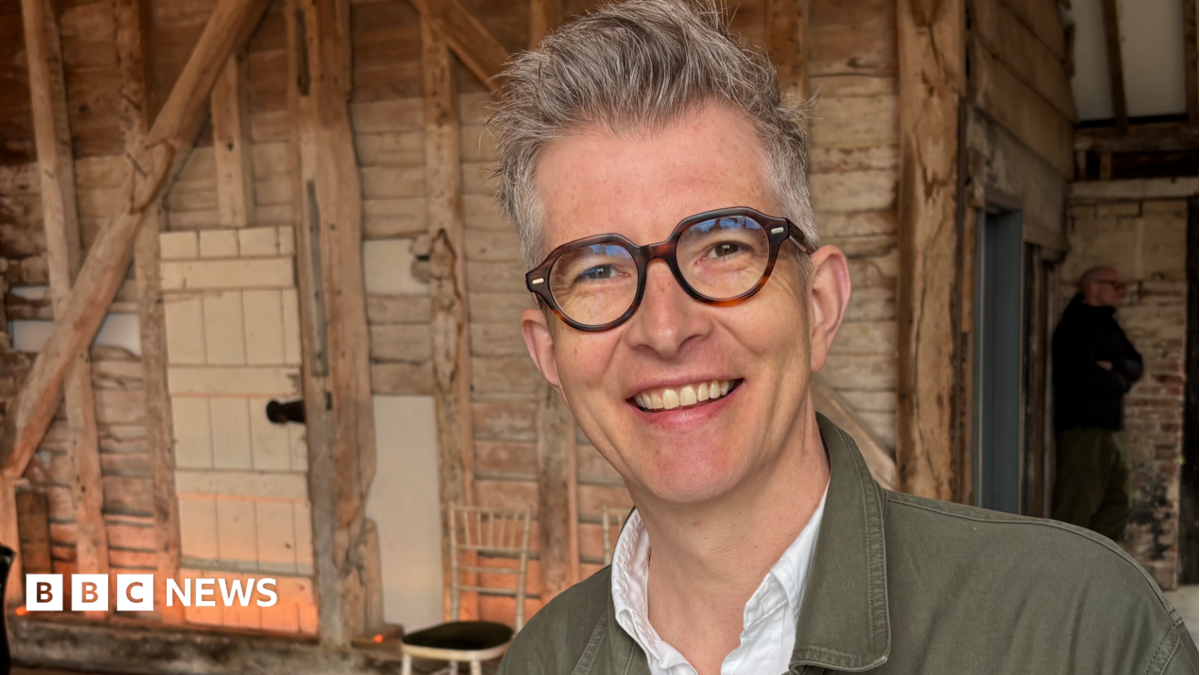 Gareth Malone smiles for the camera. He has short grey hair spiked up at the front, thick-rimmed glasses and an open-collar white shirt and khaki jean jacket.