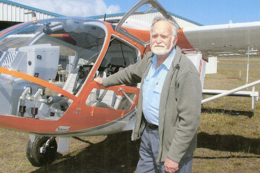 Don Adams, an older man with a white beard, stands beside a small helicopter plane. ma