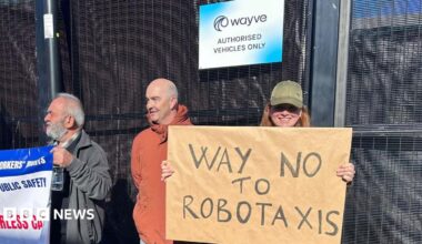 Three protesters standing outside a building with a sign that reads "Wayve Authorised Vehicles Only". One person on the left holds a union banner, a middle person in a rust-coloured jacket looks toward the right, and a person on the right smiles while holding a cardboard sign that says "Way No to Robotaxis".