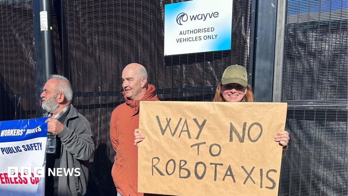 Three protesters standing outside a building with a sign that reads "Wayve Authorised Vehicles Only". One person on the left holds a union banner, a middle person in a rust-coloured jacket looks toward the right, and a person on the right smiles while holding a cardboard sign that says "Way No to Robotaxis".