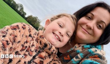 A mother and young daughter pose together in a grassy field. The young daughter wears a fluffy leopard-print fleece and her hair tied back in a ponytail. The mum wears a blue patterned fleece and has dark brown hair worn down.
