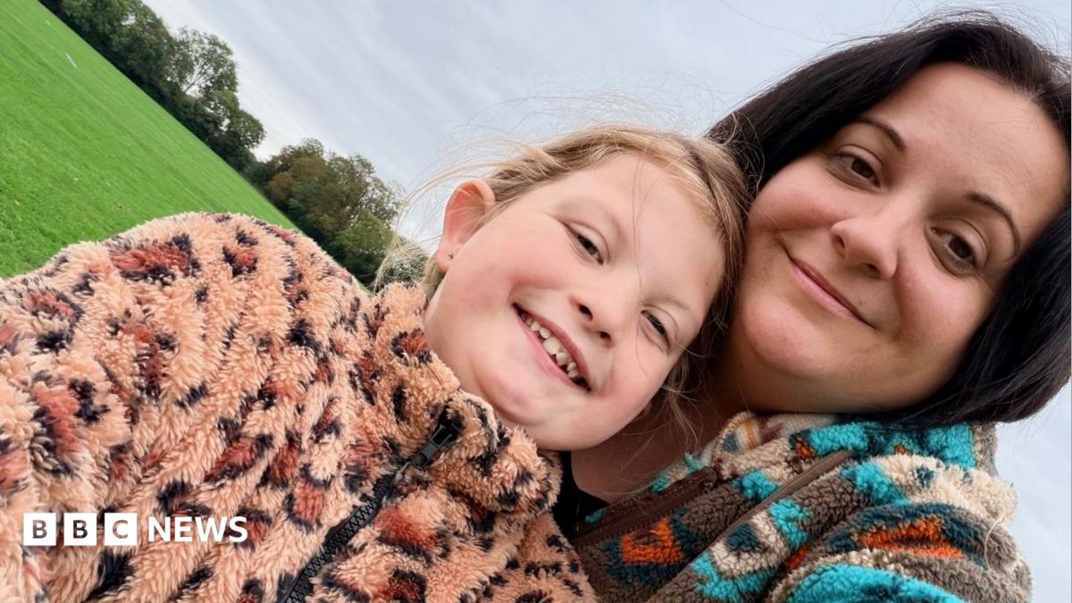 A mother and young daughter pose together in a grassy field. The young daughter wears a fluffy leopard-print fleece and her hair tied back in a ponytail. The mum wears a blue patterned fleece and has dark brown hair worn down.