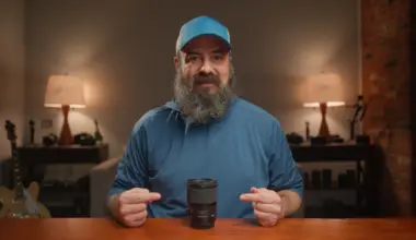 Bearded man in a blue cap and blue shirt pointing at a black camera lens standing on a wooden table