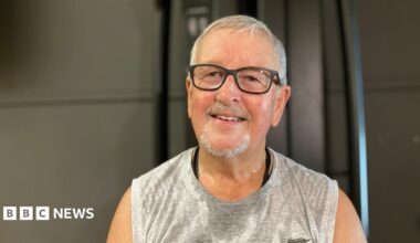 Mike James smiles at the camera as he sits in a gym. He has short grey hair, black thick-rimmed glasses and wears a grey sports vest that is damp with sweat.