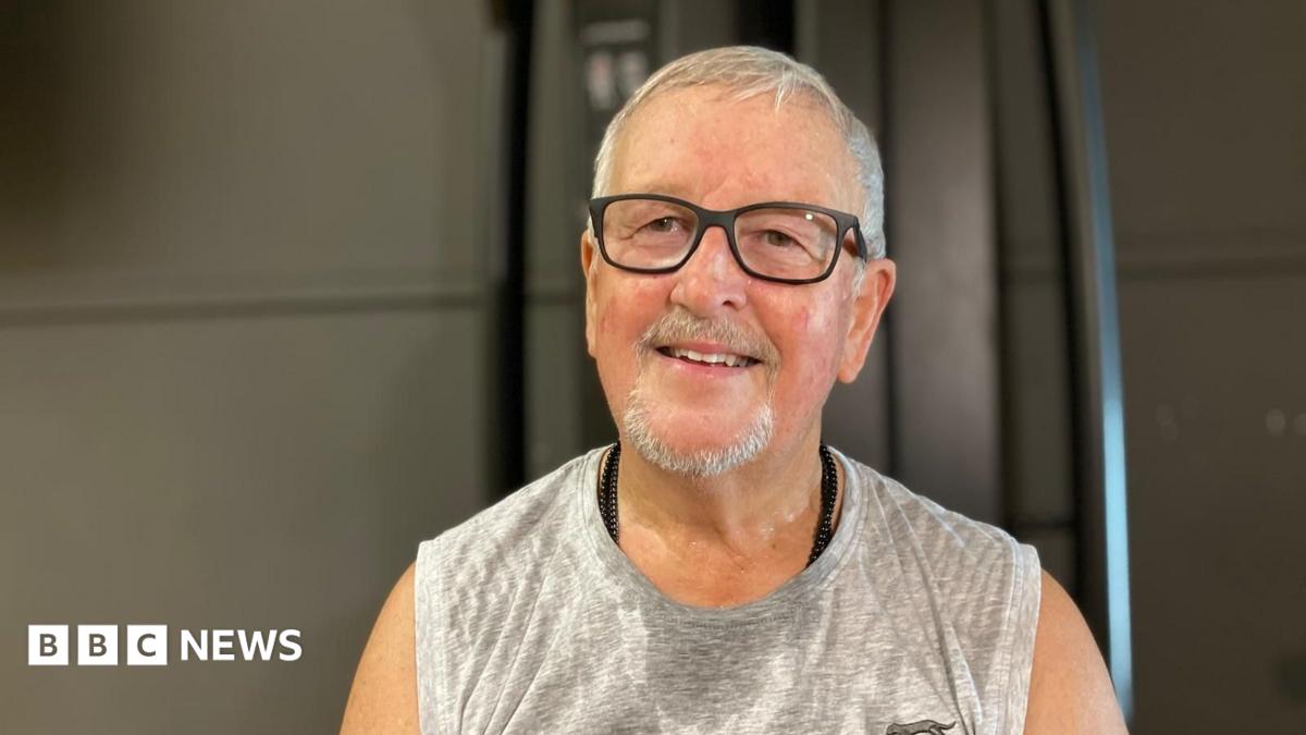 Mike James smiles at the camera as he sits in a gym. He has short grey hair, black thick-rimmed glasses and wears a grey sports vest that is damp with sweat.
