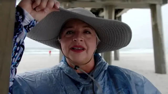 Jane speaks while holding her large hat and standing underneath a pier