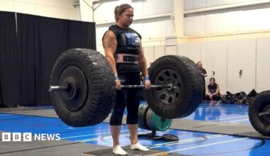 A woman wearing black sportswear and a pink weight belt lifts a barbell with large tyres on either side. She has brown hair tied back in a ponytail. In the background, a gym setting can be seen.