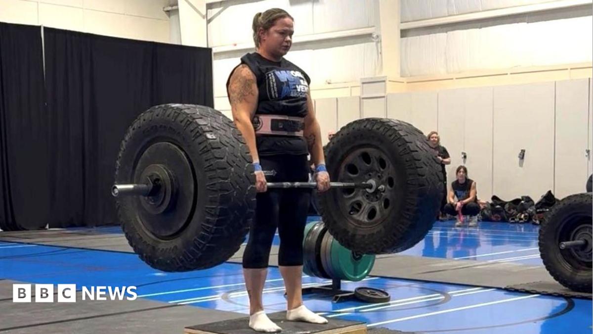 A woman wearing black sportswear and a pink weight belt lifts a barbell with large tyres on either side. She has brown hair tied back in a ponytail. In the background, a gym setting can be seen.