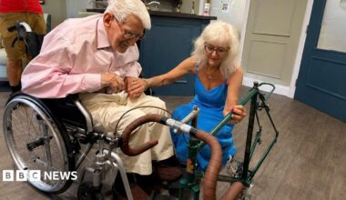 Clifford Craske and Alice Craske are in a room. He is in a wheelchair and has white hair and spectacles. He is wearing a pink shirt and cream trousers. She has long blonde hair and is wearing spectacles and a blue dress. They are both looking at the frame of a racing bicycle.