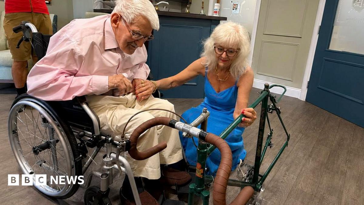 Clifford Craske and Alice Craske are in a room. He is in a wheelchair and has white hair and spectacles. He is wearing a pink shirt and cream trousers. She has long blonde hair and is wearing spectacles and a blue dress. They are both looking at the frame of a racing bicycle.