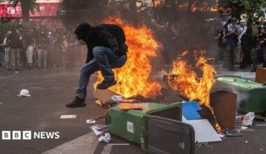 A protestor in a black hoodie jumps over a wheelie bin on fire in Paris, France.