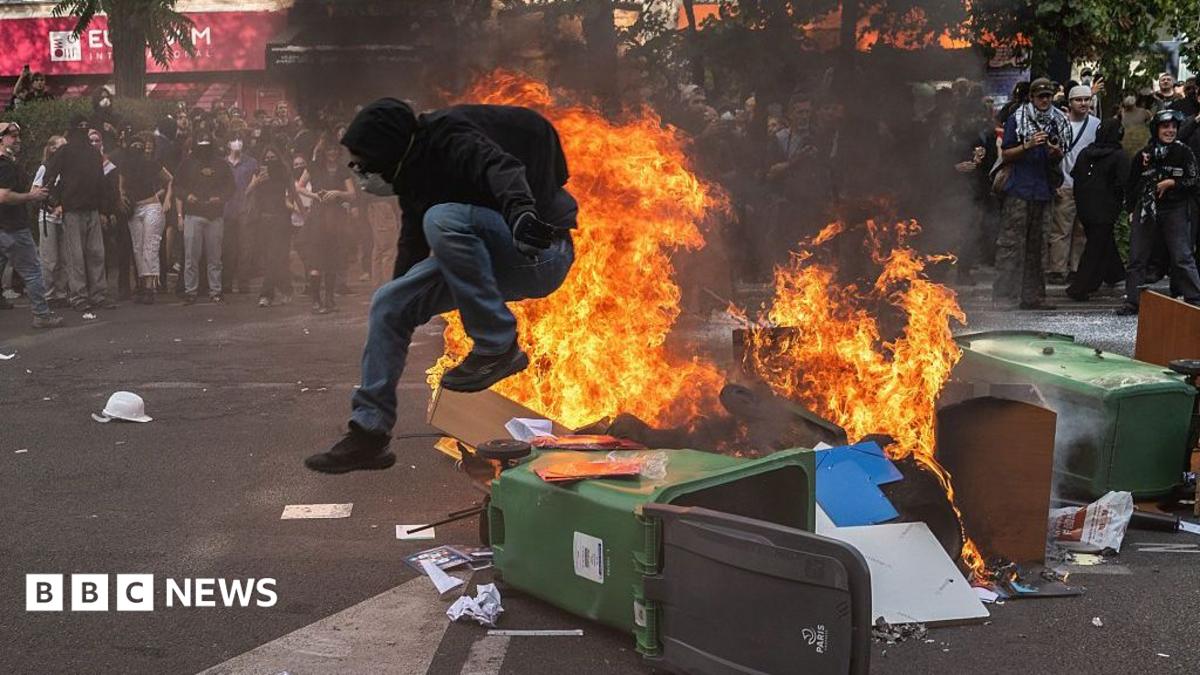 A protestor in a black hoodie jumps over a wheelie bin on fire in Paris, France.