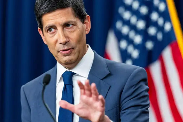 A close-up of Fed Chair Kevin Warsh in a blue suit with a US flag behind him