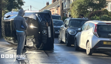 A black Mercedes is on its side in the middle of the road. There is a row of parked cars on the right hand side and a person in a grey sports tracksuit is standing in front of the car and is speaking into his phone.