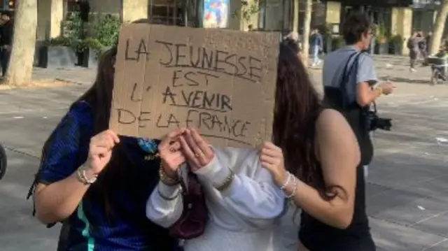 Three young women hide their faces behind a cardboard sign with the message: "Youth is the future of France" written in French