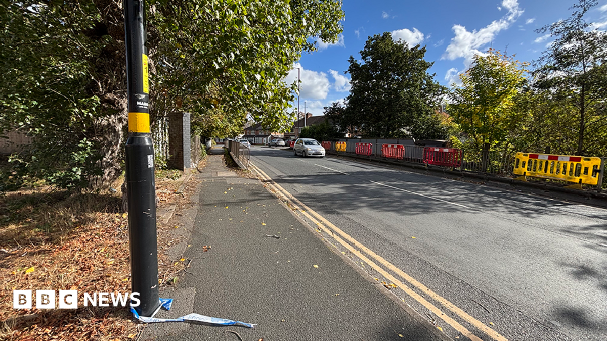 Some railings either side of a road with a lamp-post on the left in the foreground. The remains of some police tape are tied to the base of the lamp-post