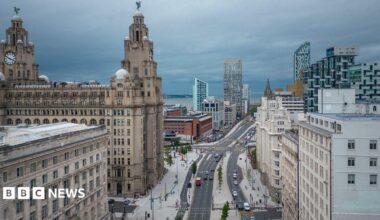 Elevated view of Liverpool city centre showing the Royal Liver Building on the left, a wide multi-lane road running through the centre, and a mix of historic and modern buildings on either side. Cars travel along a roadway lined with young trees and pedestrian walkways, while contemporary high-rise developments form part of the skyline under a cloudy grey sky.