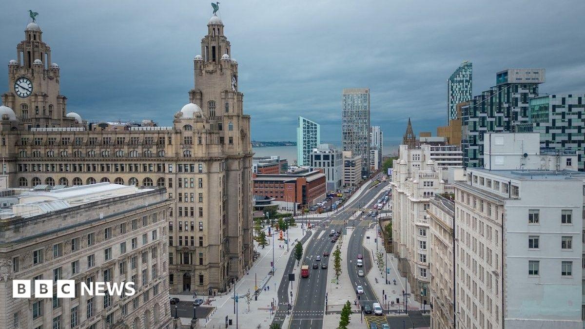 Elevated view of Liverpool city centre showing the Royal Liver Building on the left, a wide multi-lane road running through the centre, and a mix of historic and modern buildings on either side. Cars travel along a roadway lined with young trees and pedestrian walkways, while contemporary high-rise developments form part of the skyline under a cloudy grey sky.