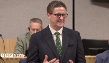 A man in a navy suit, green tie, and glasses, stands up and speaks inside a formal-looking room.
