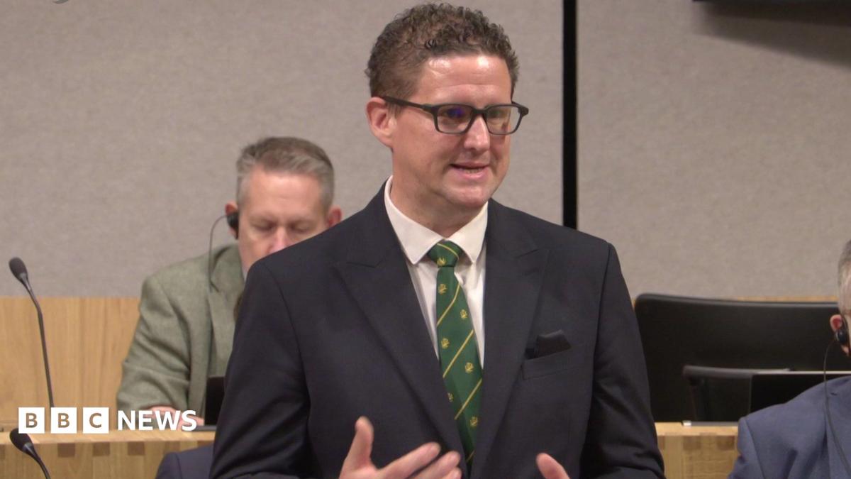 A man in a navy suit, green tie, and glasses, stands up and speaks inside a formal-looking room.