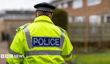 A police officer is walking away from the camera. He wears a yellow high-vis coat, which has silver reflective panels down the sides and a rectangular logo along the centre which reads 'POLICE' on a blue background. He also wears a black cap with a black and white check panel around the head. The background is blurred but shows a housing estate.