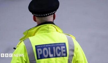 The back of a Greater Manchester Police officer who is wearing a fluorescent yellow high-visibility jacket and black police cap, with a large blue and white “POLICE” badge on the back, standing against a blurred grey background.