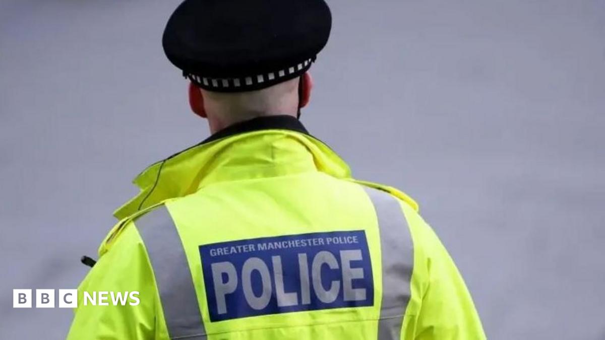 The back of a Greater Manchester Police officer who is wearing a fluorescent yellow high-visibility jacket and black police cap, with a large blue and white “POLICE” badge on the back, standing against a blurred grey background.