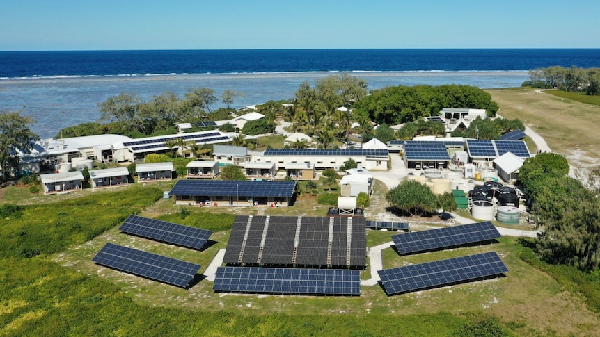 A vast number of solar panels in several rows in the foreground, with a cluster of small white resort buildings by the ocean.