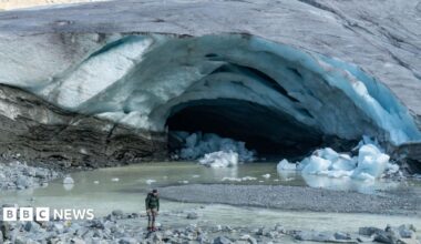 The front of a glacier, where a large channel - which looks a bit like a cave or an arch - has eaten away at the ice. A river of meltwater now meanders out of the front of the glacier. There are also chunks of ice on the ground having broken off the glacier. A man, dressed in mountaineering clothing, stands in front of the glacier for scale, which shows how big the channel or cave is.