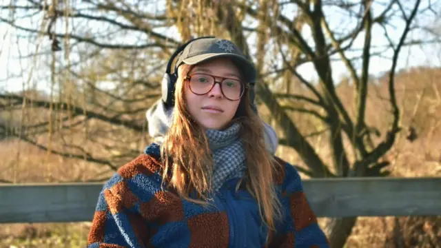 Erin is pictured wearing glasses, headphones, a cap and a fleece jacket, standing outdoors in front of leafless trees. Warm low sunlight illuminates the scene, with a wooden fence and blurred natural landscape in the background.
