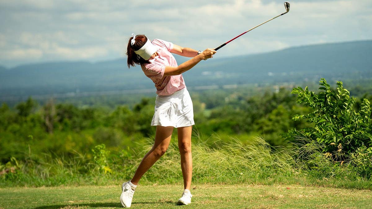 Woman in pink shirt and white skirt swings golf club on course.