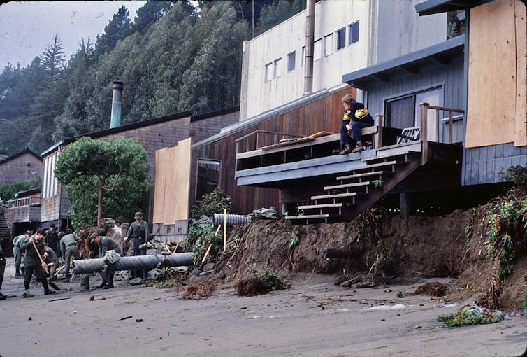 A person sits on a porch with stairs that end stop about 8 feet above the beach.