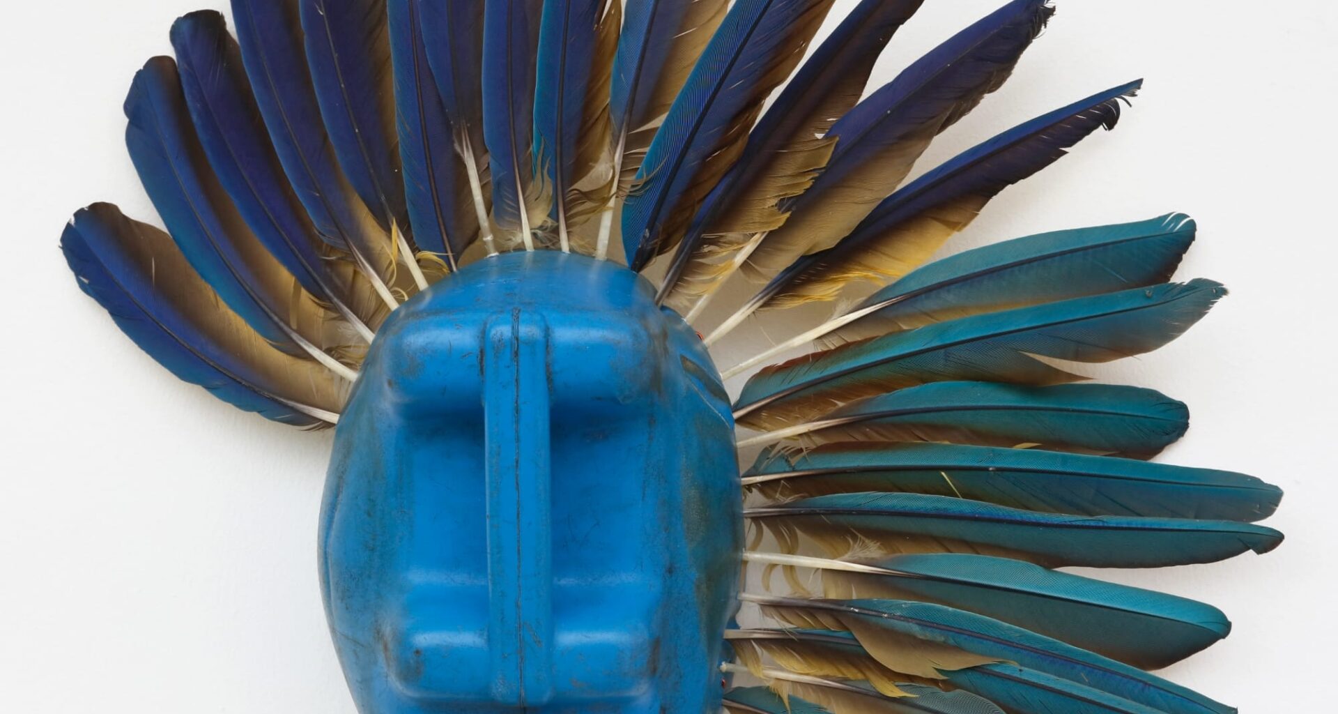 an abstract mask-like sculpture made of a blue plastic jug and blue and tan feathers