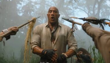 large bald very muscular man in tight shirt standing open mouthed while surrounded by jungle natives brandishing spears