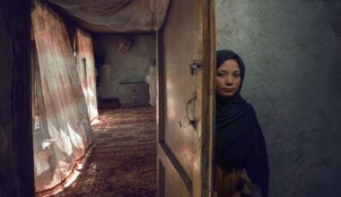 A young girl in a dark headscarf peers out from behind a half-open wooden door with a metal bolt, standing against a dim grey wall, while beyond the door a sunlit room with patterned curtains