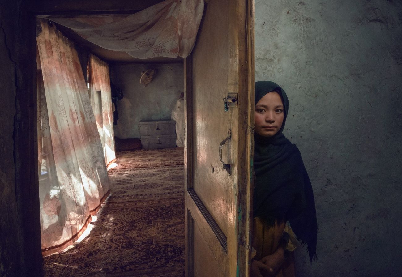 A young girl in a dark headscarf peers out from behind a half-open wooden door with a metal bolt, standing against a dim grey wall, while beyond the door a sunlit room with patterned curtains