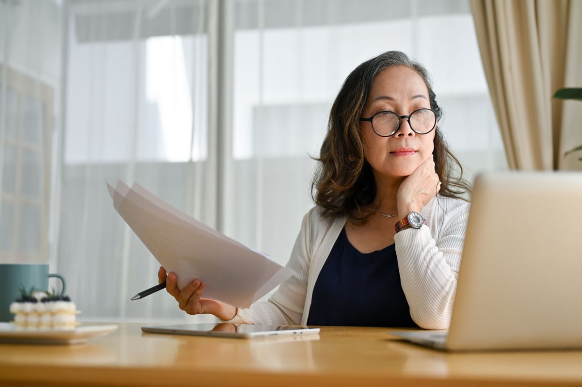 Mature woman in glasses reviewing documents while working at a laptop in a bright home office