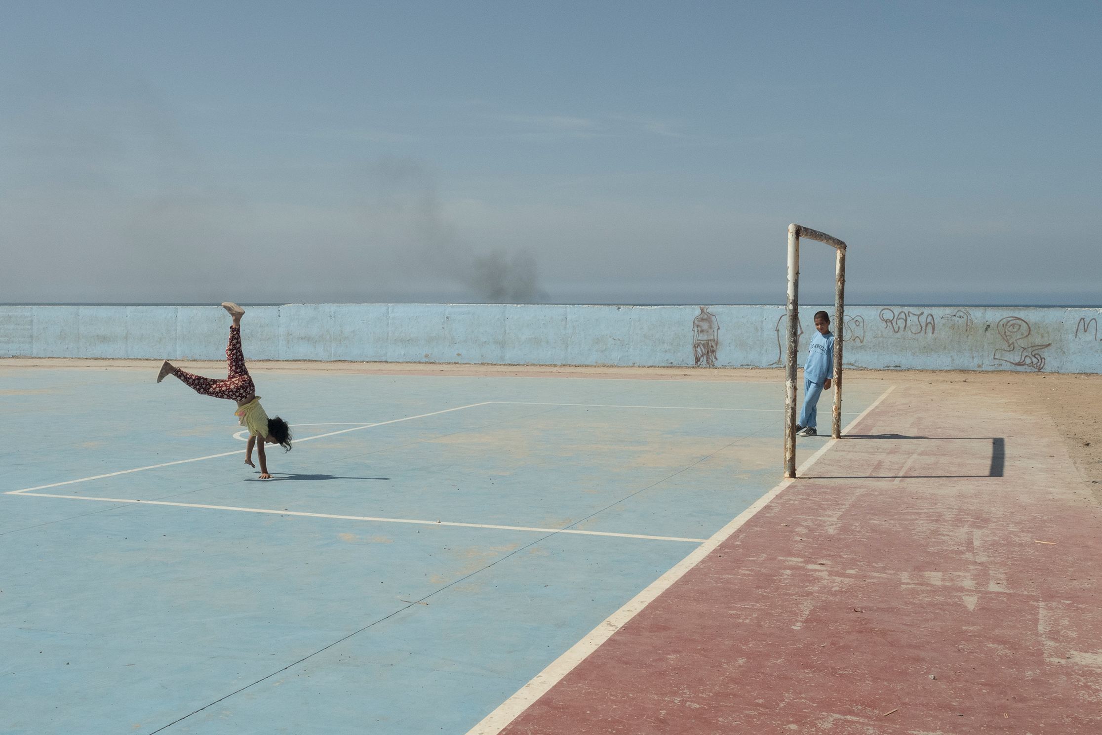 Child doing a handstand on a faded outdoor basketball court while another child stands by the goal