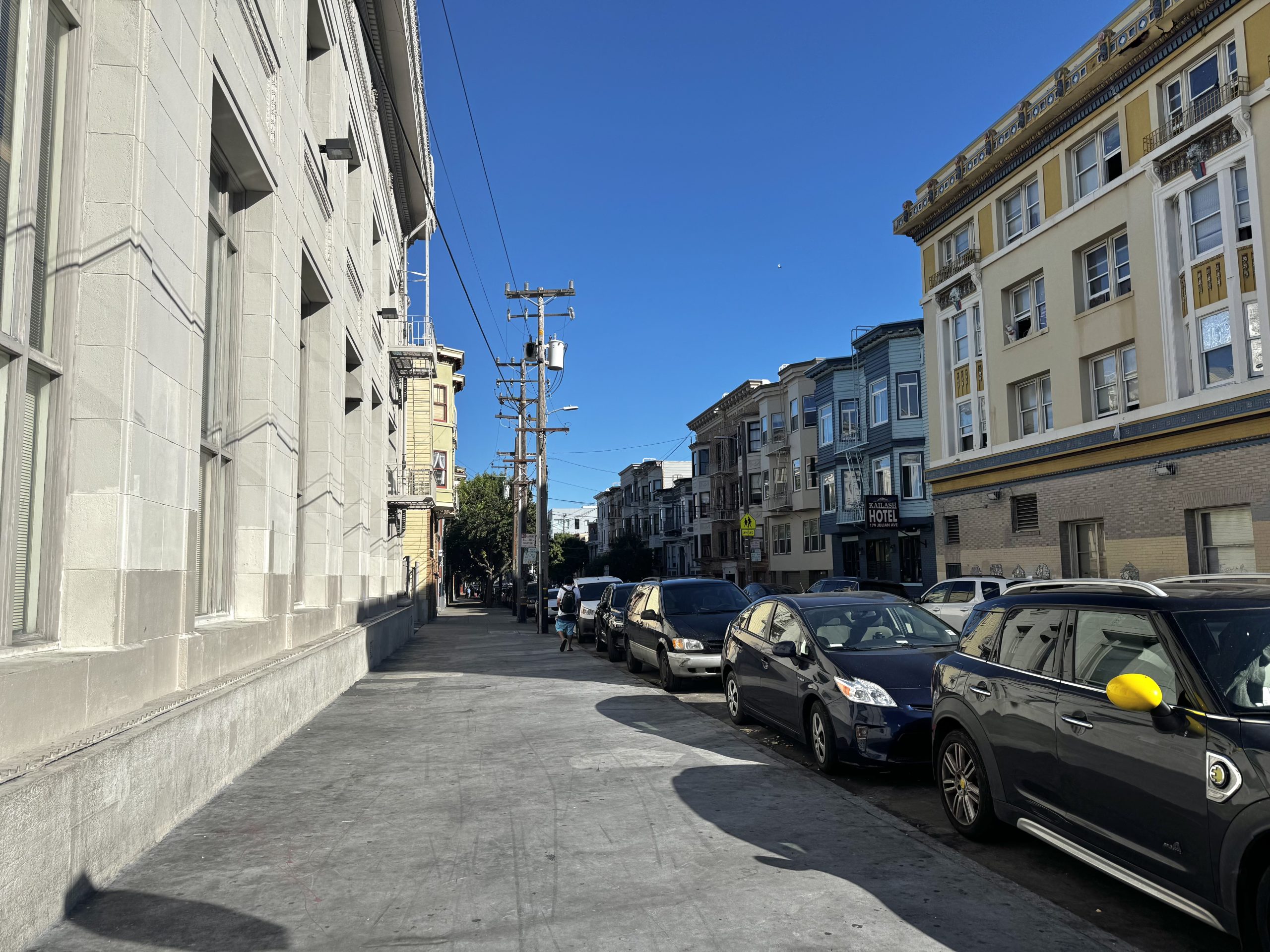 A city street lined with parked cars and multi-story buildings under a clear blue sky.