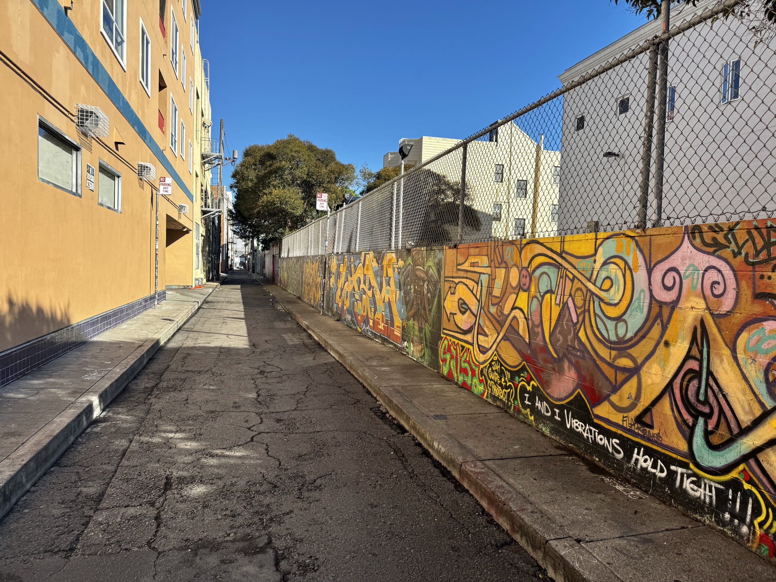 A narrow urban alley with colorful graffiti on a wall, a chain-link fence, cracked pavement, and buildings under a clear blue sky.