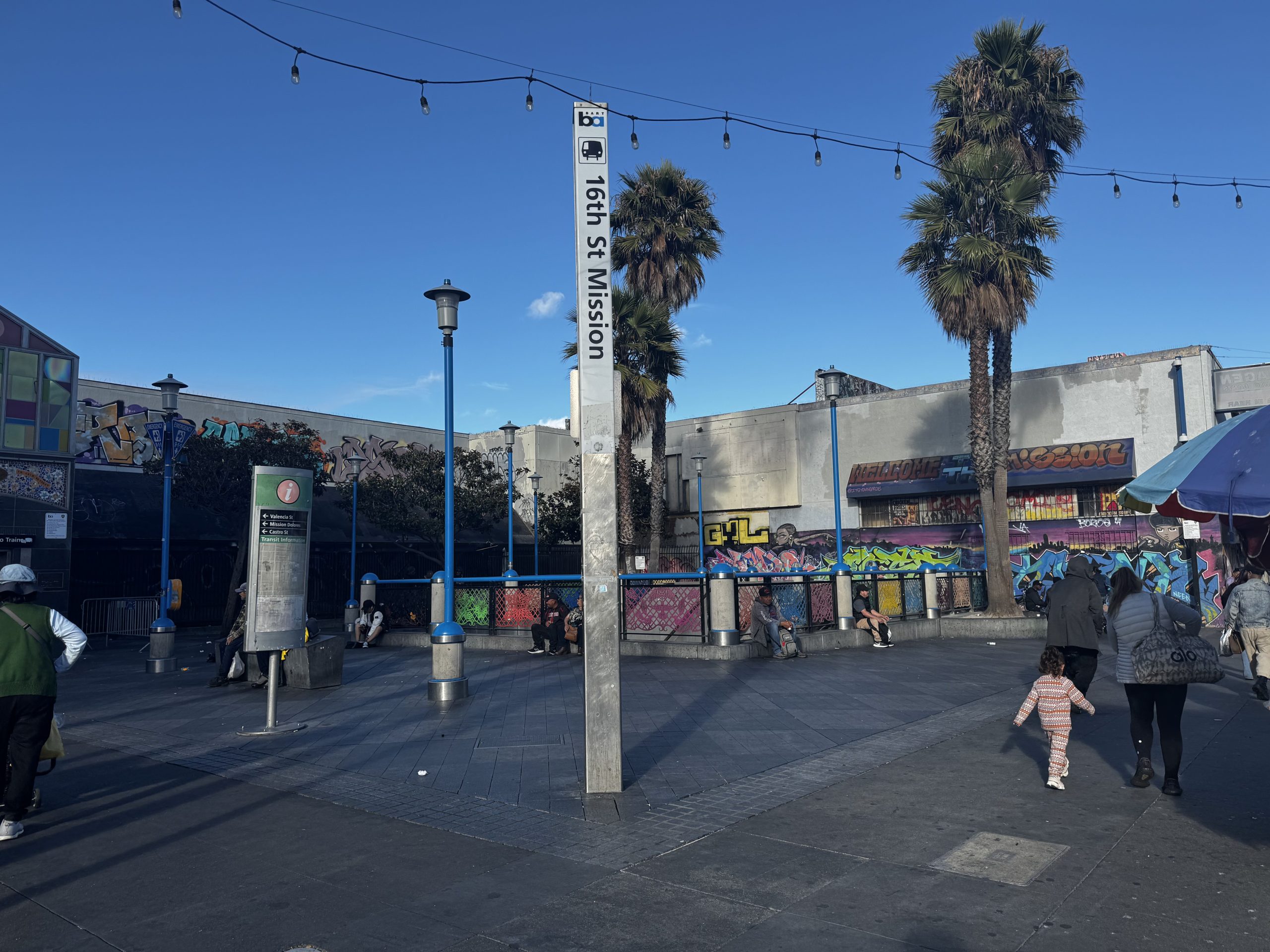 16th St Mission BART plaza with people walking, palm trees, string lights, and colorful graffiti murals on surrounding buildings under a clear blue sky.