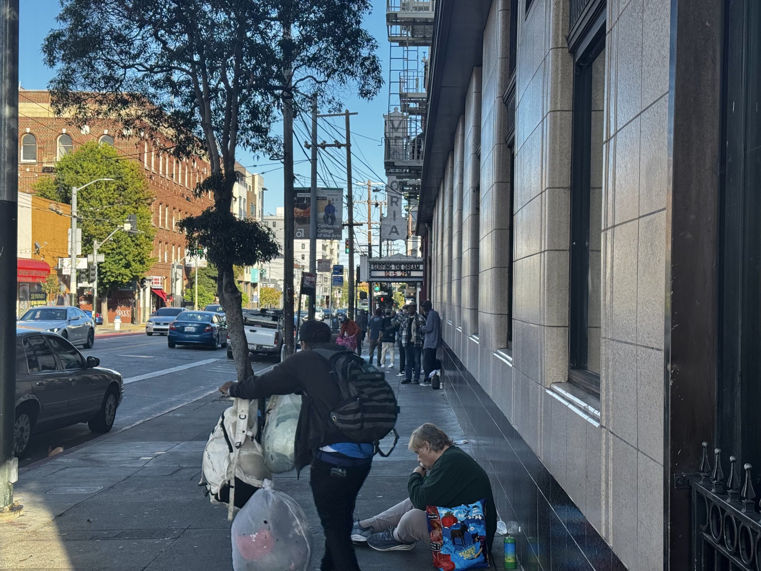 People walk and sit along a city sidewalk next to a building; one person pushes a cart while another sits with bags near the wall. Cars are parked on the street.