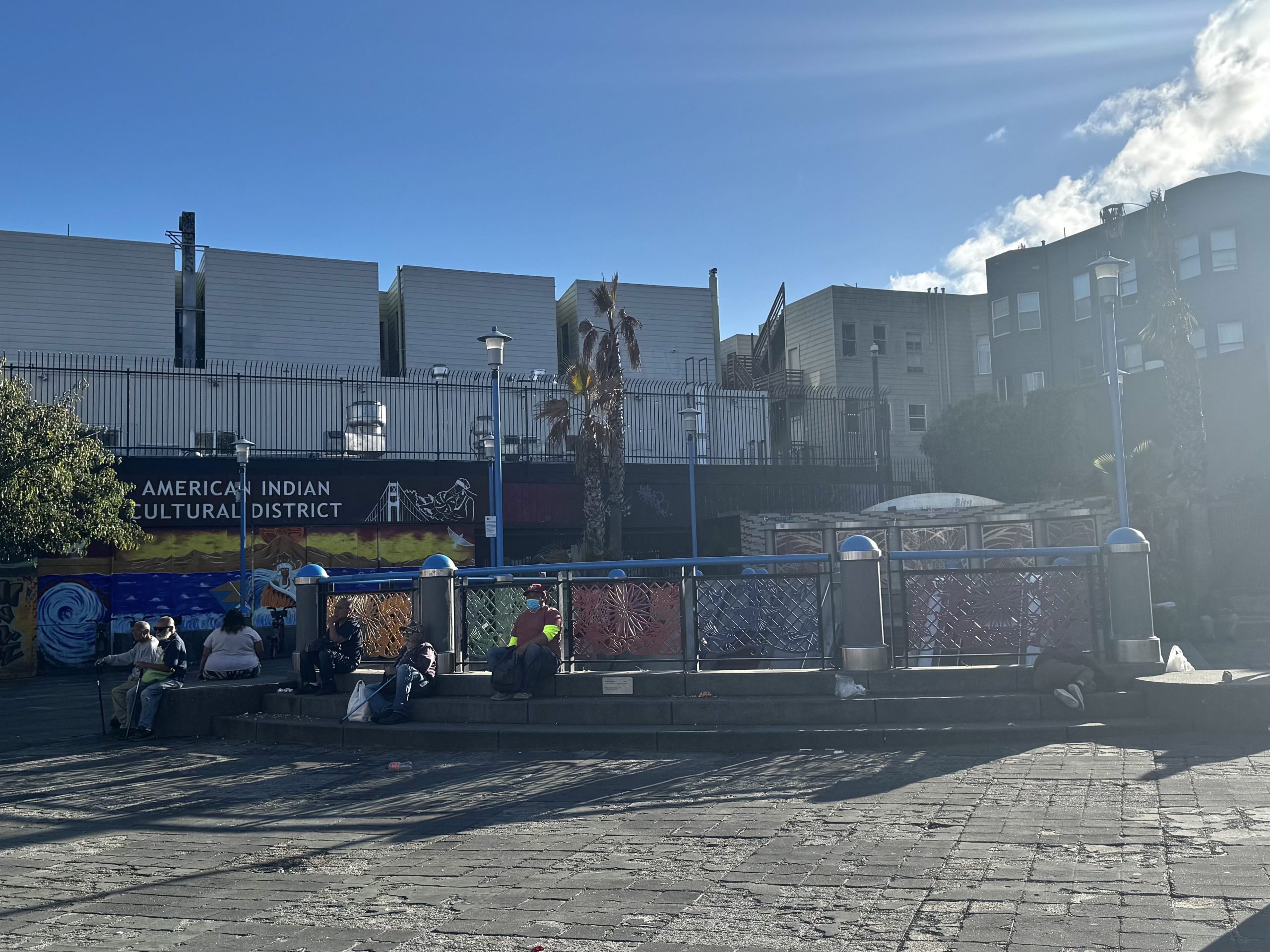 A group of people sits on benches in an urban plaza near a mural that reads "American Indian Cultural District" on a sunny day.