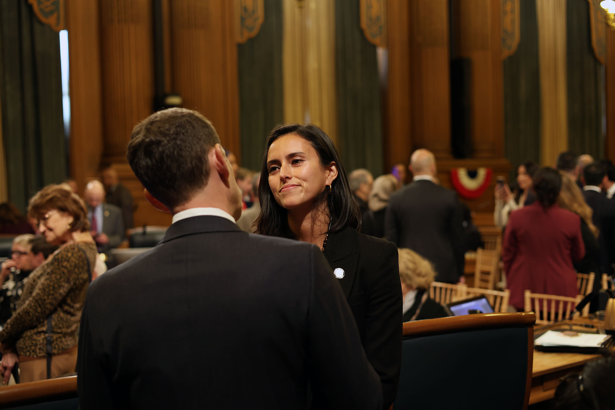 A woman and a man engage in conversation in a formal setting with people seated and standing in the background.