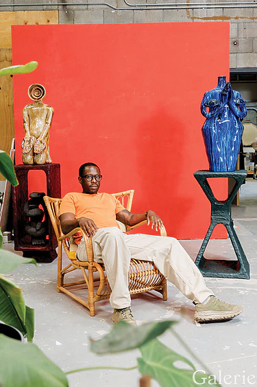 Man sitting in a wicker chair in an art studio with sculptures and a red wall in the background