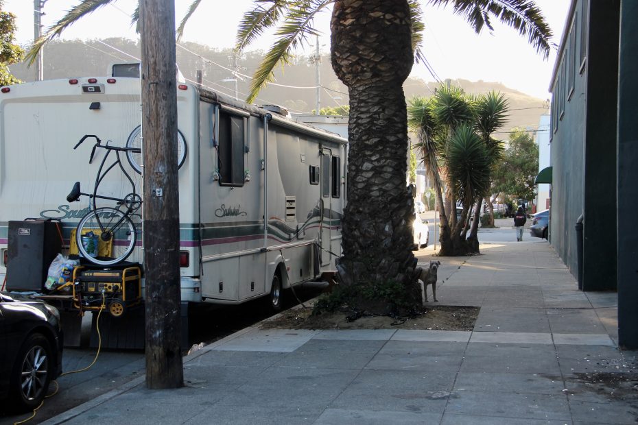 A large RV with a bike attached to the back is parked on a city street next to a palm tree and sidewalk in daylight.