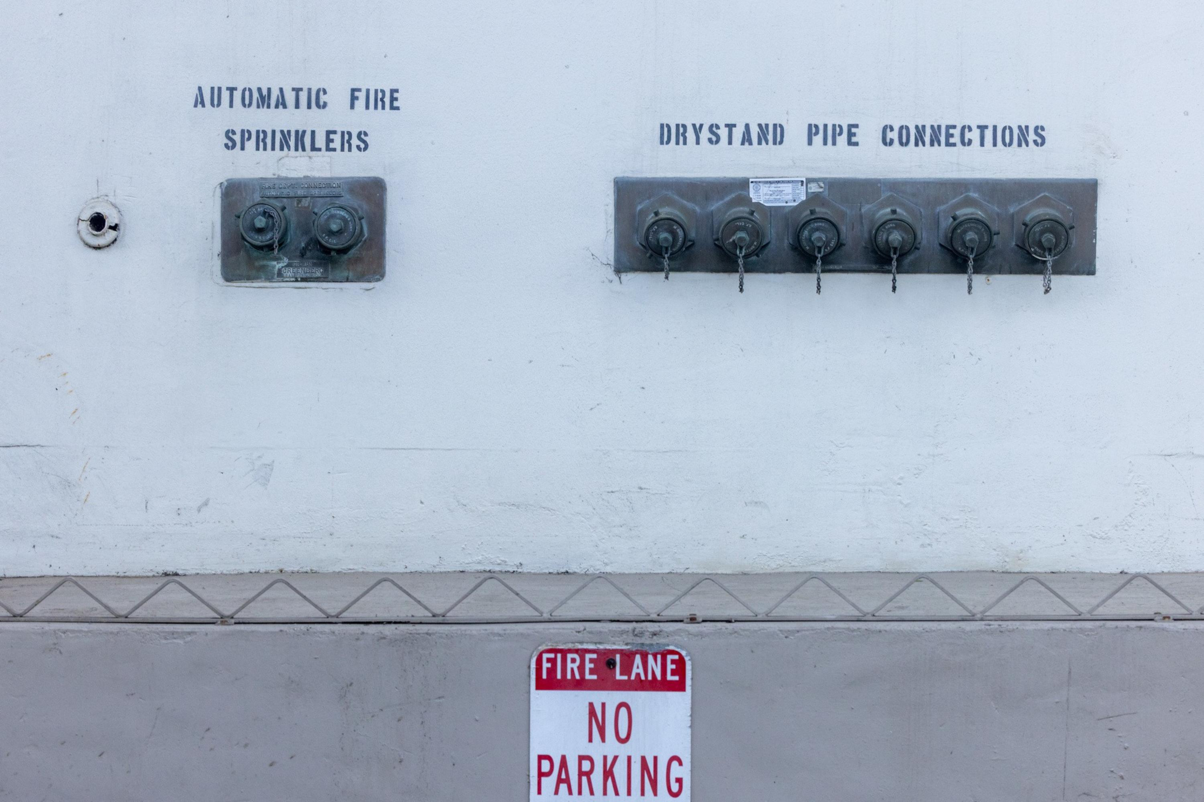 Wall-mounted fire safety equipment labeled “Automatic Fire Sprinklers” with two valves and “Drystand Pipe Connections” with six valves, above a “Fire Lane No Parking” sign.