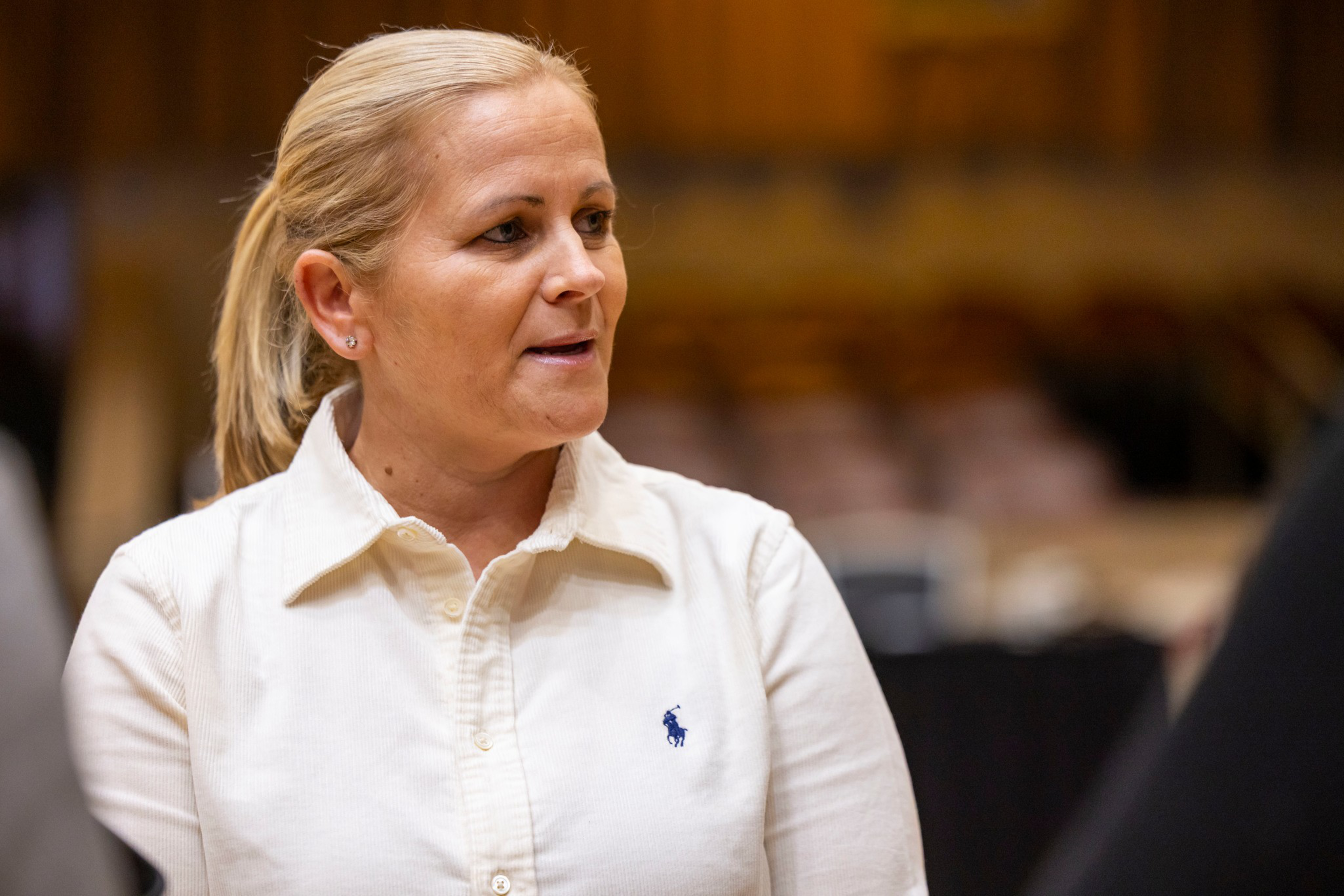 A woman with blonde hair tied back wears a white collared shirt with a small blue logo, speaking or listening attentively in a softly blurred indoor setting.